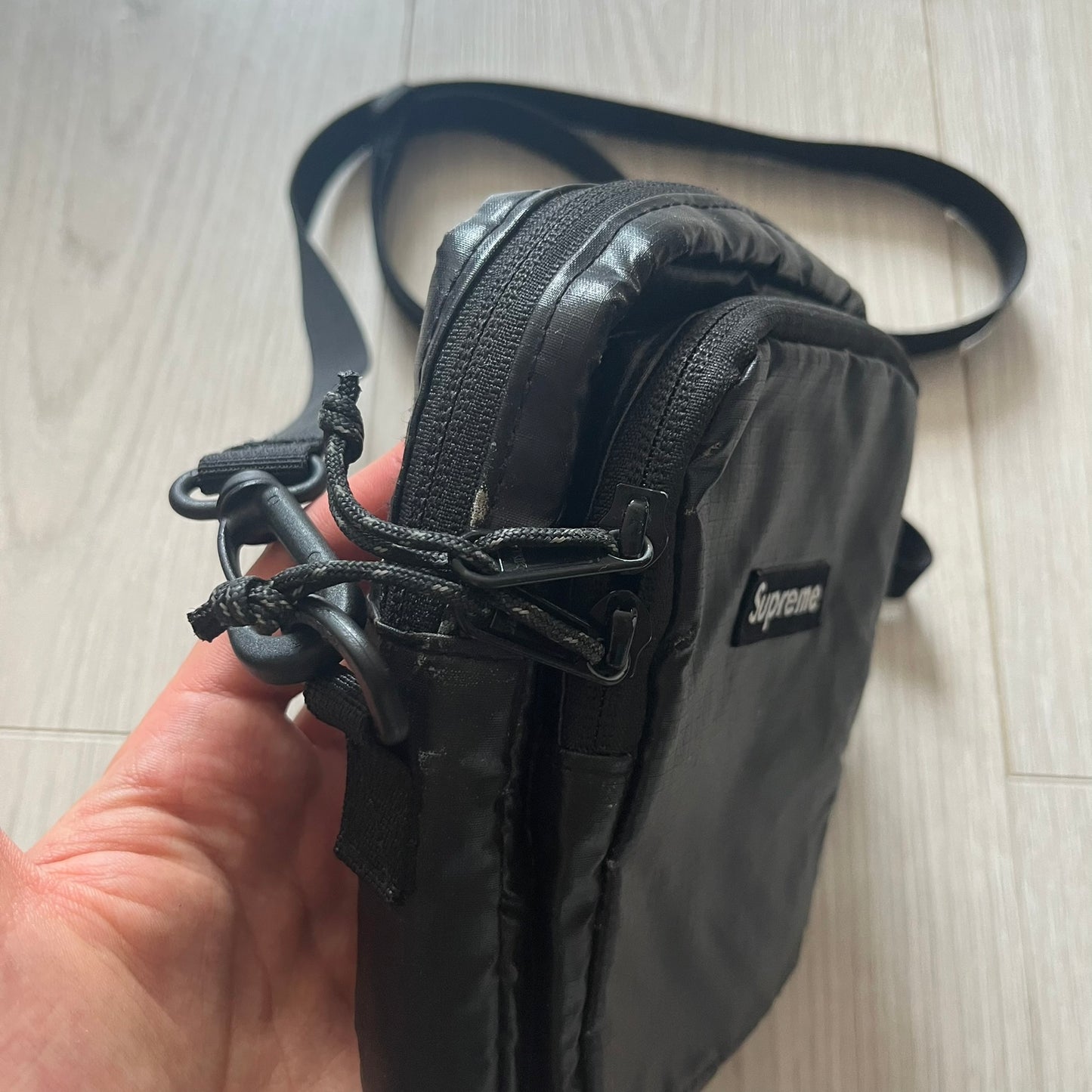 Black Supreme bag held by a hand on a light wooden floor.