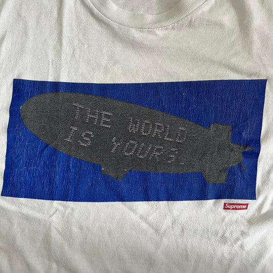 White t-shirt with a blue and gray graphic design featuring 'The World is Yours' text and a Supreme logo.