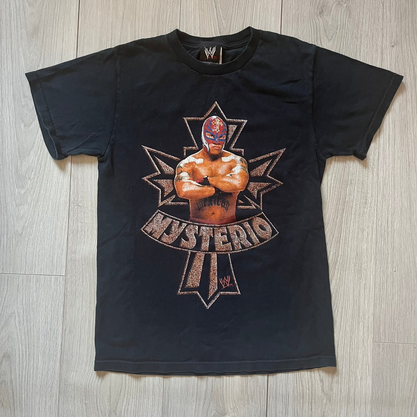 Black t-shirt with a graphic of a wrestler and 'Mysterio' text on a wooden floor.