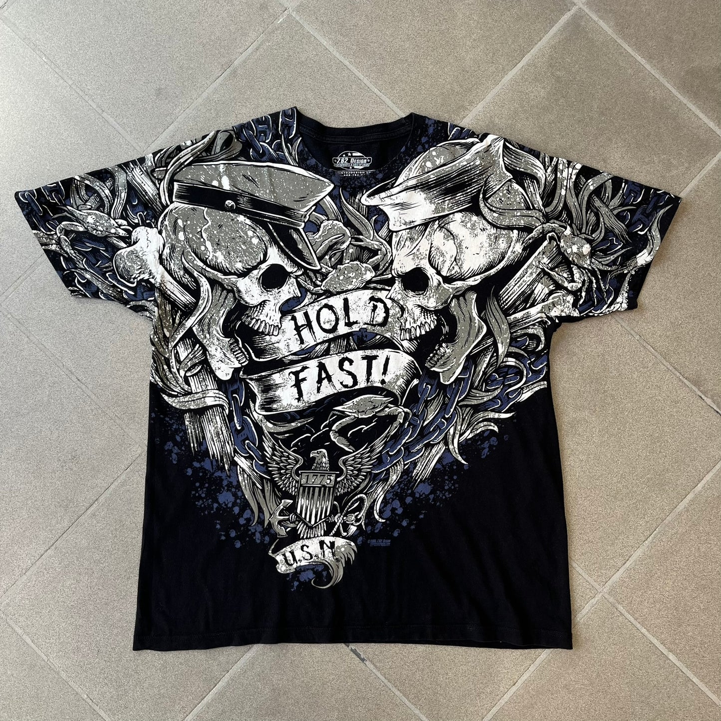 7.62 Design US Navy Black Tee features large front and back graphics in black, white, grey, and navy. The front has two skulls, bones, and US Navy graphics covering most of the shirt. The back has a smaller US Navy logo graphic.
