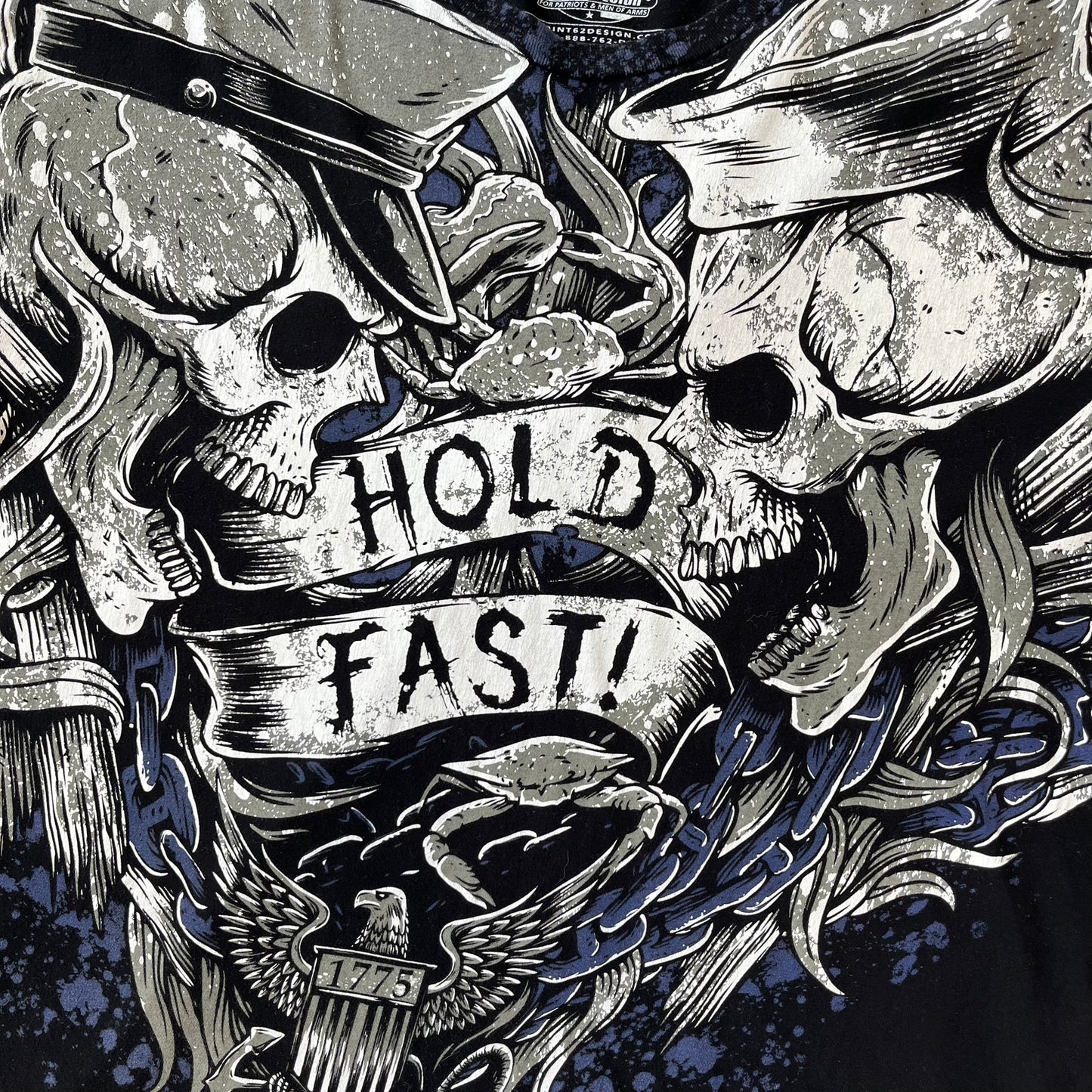 7.62 Design US Navy Black Tee features large front and back graphics in black, white, grey, and navy. The front has two skulls, bones, and US Navy graphics covering most of the shirt. The back has a smaller US Navy logo graphic.
