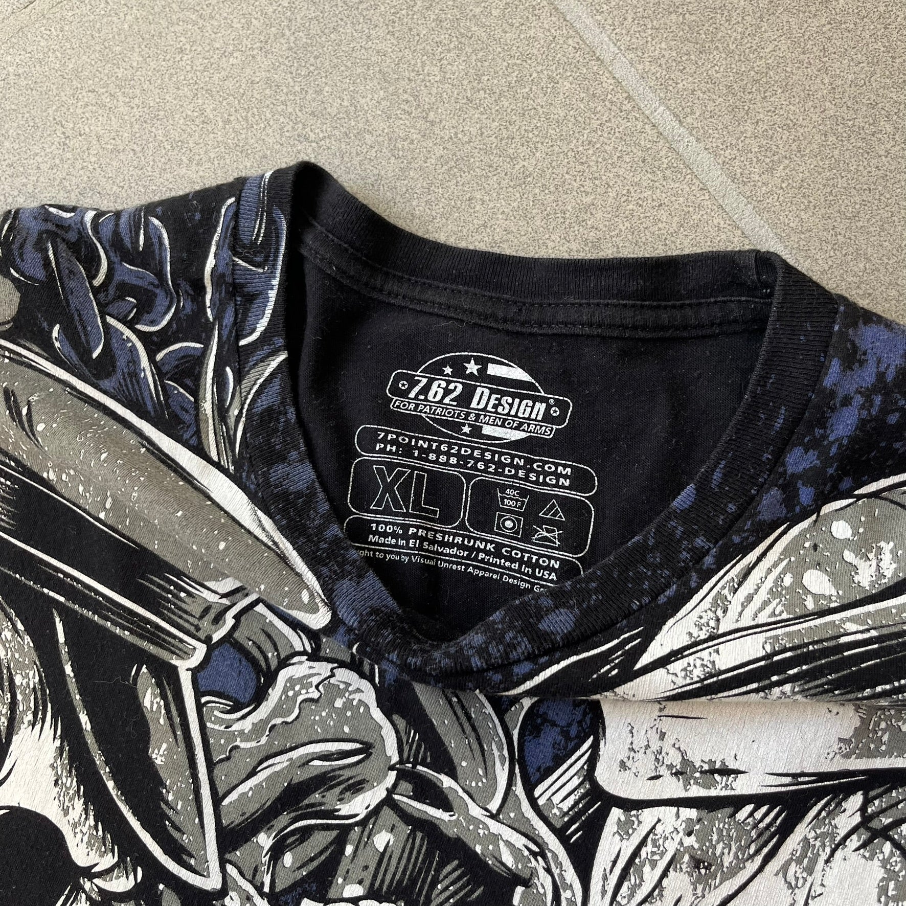 7.62 Design US Navy Black Tee features large front and back graphics in black, white, grey, and navy. The front has two skulls, bones, and US Navy graphics covering most of the shirt. The back has a smaller US Navy logo graphic.

