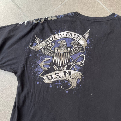 7.62 Design US Navy Black Tee features large front and back graphics in black, white, grey, and navy. The front has two skulls, bones, and US Navy graphics covering most of the shirt. The back has a smaller US Navy logo graphic.
