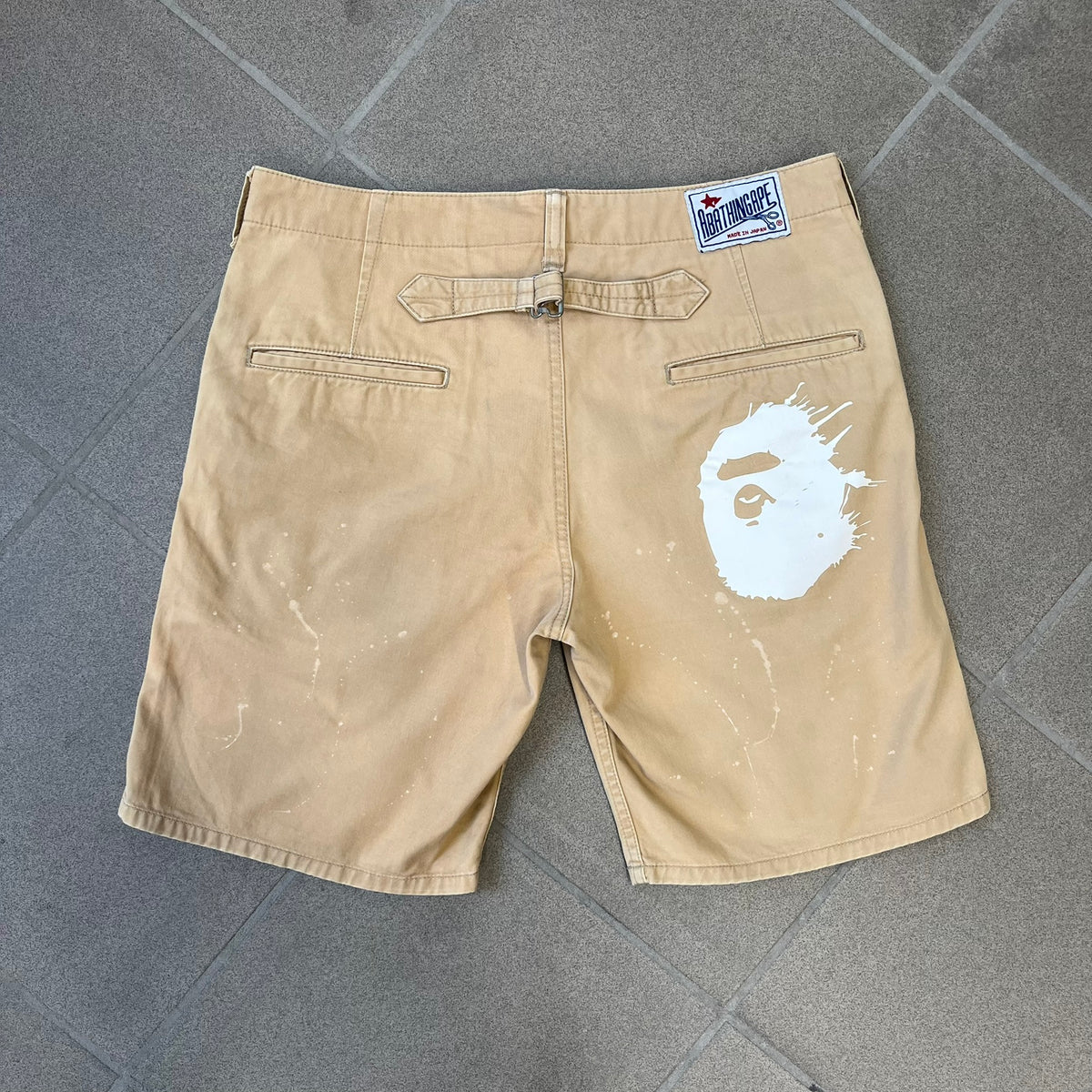 A Bathing Ape 2000’s Paint Splatter Beige Chino Shorts are beige BAPE shorts made in Japan, with light white paint splatters across the front and back. A white Bape head on the back has the same painted splatter effect. The shorts have front and back pockets, a Bathing Ape patch on the back, and branded buttons.