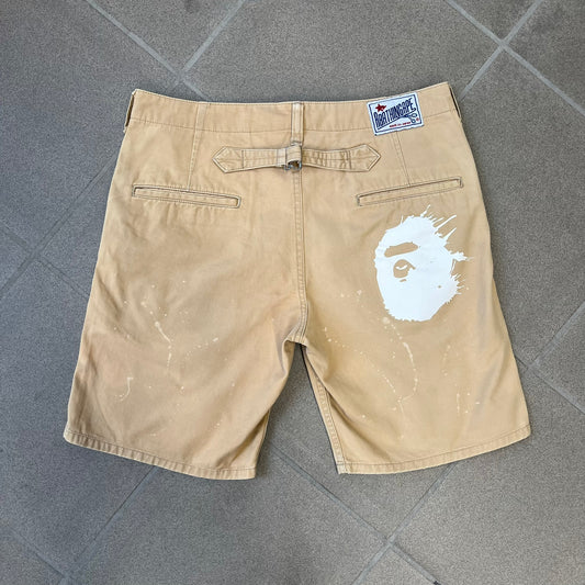 A Bathing Ape 2000’s Paint Splatter Beige Chino Shorts are beige BAPE shorts made in Japan, with light white paint splatters across the front and back. A white Bape head on the back has the same painted splatter effect. The shorts have front and back pockets, a Bathing Ape patch on the back, and branded buttons.