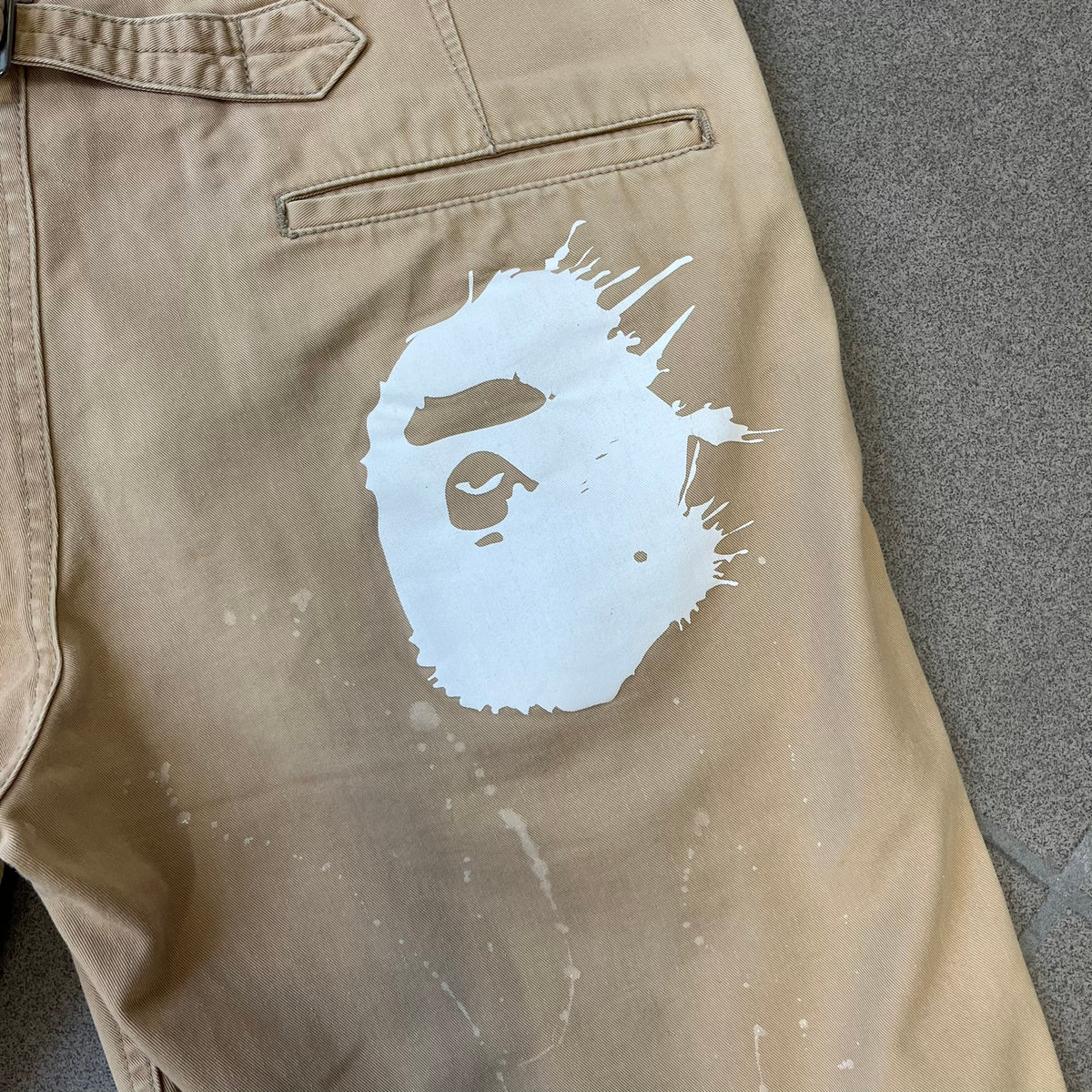 A Bathing Ape 2000’s Paint Splatter Beige Chino Shorts are beige BAPE shorts made in Japan, with light white paint splatters across the front and back. A white Bape head on the back has the same painted splatter effect. The shorts have front and back pockets, a Bathing Ape patch on the back, and branded buttons.