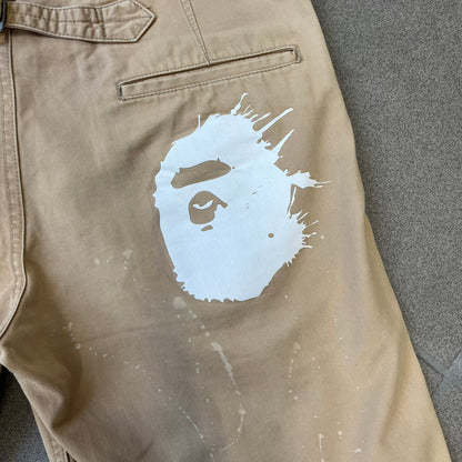 A Bathing Ape 2000’s Paint Splatter Beige Chino Shorts are beige BAPE shorts made in Japan, with light white paint splatters across the front and back. A white Bape head on the back has the same painted splatter effect. The shorts have front and back pockets, a Bathing Ape patch on the back, and branded buttons.