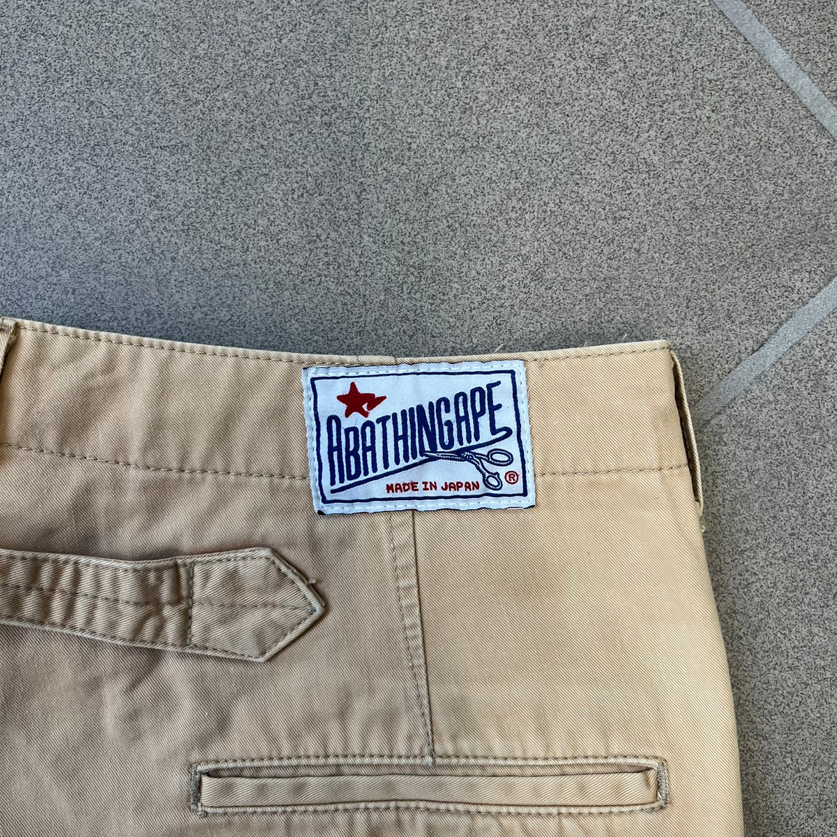 A Bathing Ape 2000’s Paint Splatter Beige Chino Shorts are beige BAPE shorts made in Japan, with light white paint splatters across the front and back. A white Bape head on the back has the same painted splatter effect. The shorts have front and back pockets, a Bathing Ape patch on the back, and branded buttons.