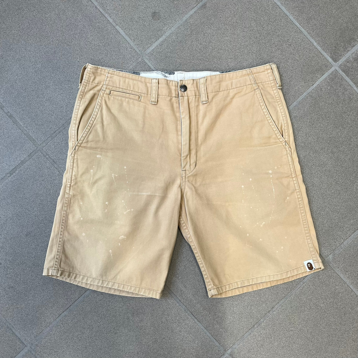 A Bathing Ape 2000’s Paint Splatter Beige Chino Shorts are beige BAPE shorts made in Japan, with light white paint splatters across the front and back. A white Bape head on the back has the same painted splatter effect. The shorts have front and back pockets, a Bathing Ape patch on the back, and branded buttons.