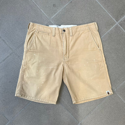 A Bathing Ape 2000’s Paint Splatter Beige Chino Shorts are beige BAPE shorts made in Japan, with light white paint splatters across the front and back. A white Bape head on the back has the same painted splatter effect. The shorts have front and back pockets, a Bathing Ape patch on the back, and branded buttons.