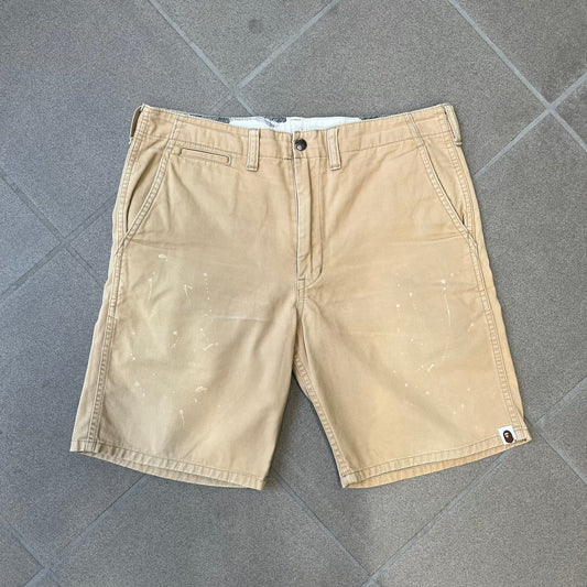 A Bathing Ape 2000’s Paint Splatter Beige Chino Shorts are beige BAPE shorts made in Japan, with light white paint splatters across the front and back. A white Bape head on the back has the same painted splatter effect. The shorts have front and back pockets, a Bathing Ape patch on the back, and branded buttons.