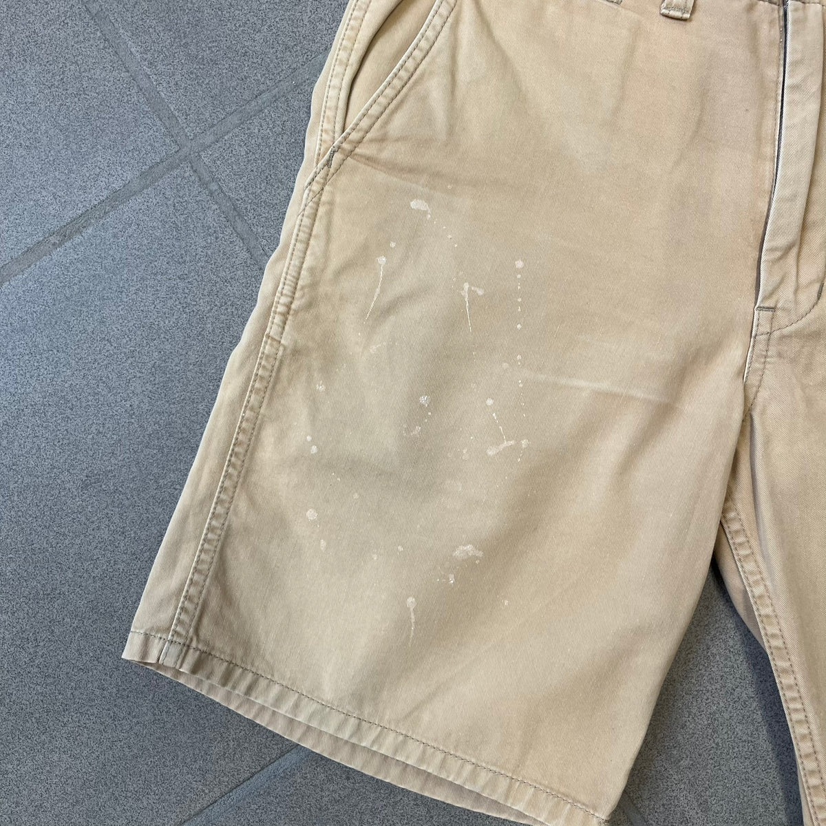 A Bathing Ape 2000’s Paint Splatter Beige Chino Shorts are beige BAPE shorts made in Japan, with light white paint splatters across the front and back. A white Bape head on the back has the same painted splatter effect. The shorts have front and back pockets, a Bathing Ape patch on the back, and branded buttons.