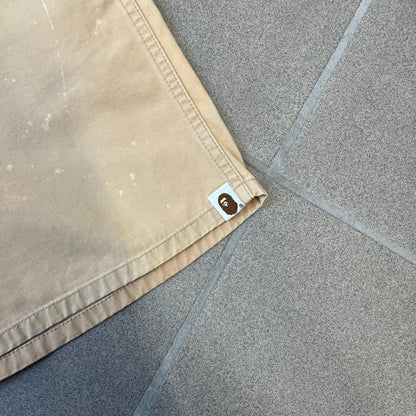 A Bathing Ape 2000’s Paint Splatter Beige Chino Shorts are beige BAPE shorts made in Japan, with light white paint splatters across the front and back. A white Bape head on the back has the same painted splatter effect. The shorts have front and back pockets, a Bathing Ape patch on the back, and branded buttons.