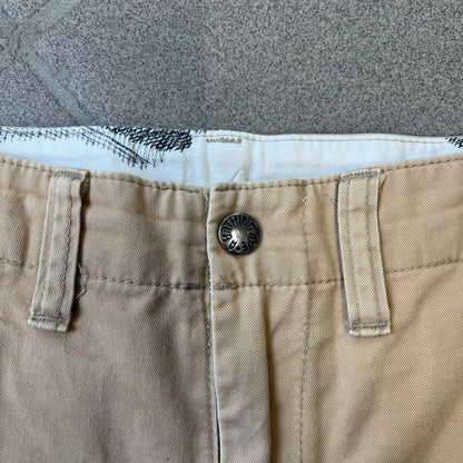 A Bathing Ape 2000’s Paint Splatter Beige Chino Shorts are beige BAPE shorts made in Japan, with light white paint splatters across the front and back. A white Bape head on the back has the same painted splatter effect. The shorts have front and back pockets, a Bathing Ape patch on the back, and branded buttons.
