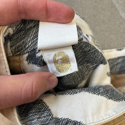 A Bathing Ape 2000’s Paint Splatter Beige Chino Shorts are beige BAPE shorts made in Japan, with light white paint splatters across the front and back. A white Bape head on the back has the same painted splatter effect. The shorts have front and back pockets, a Bathing Ape patch on the back, and branded buttons.