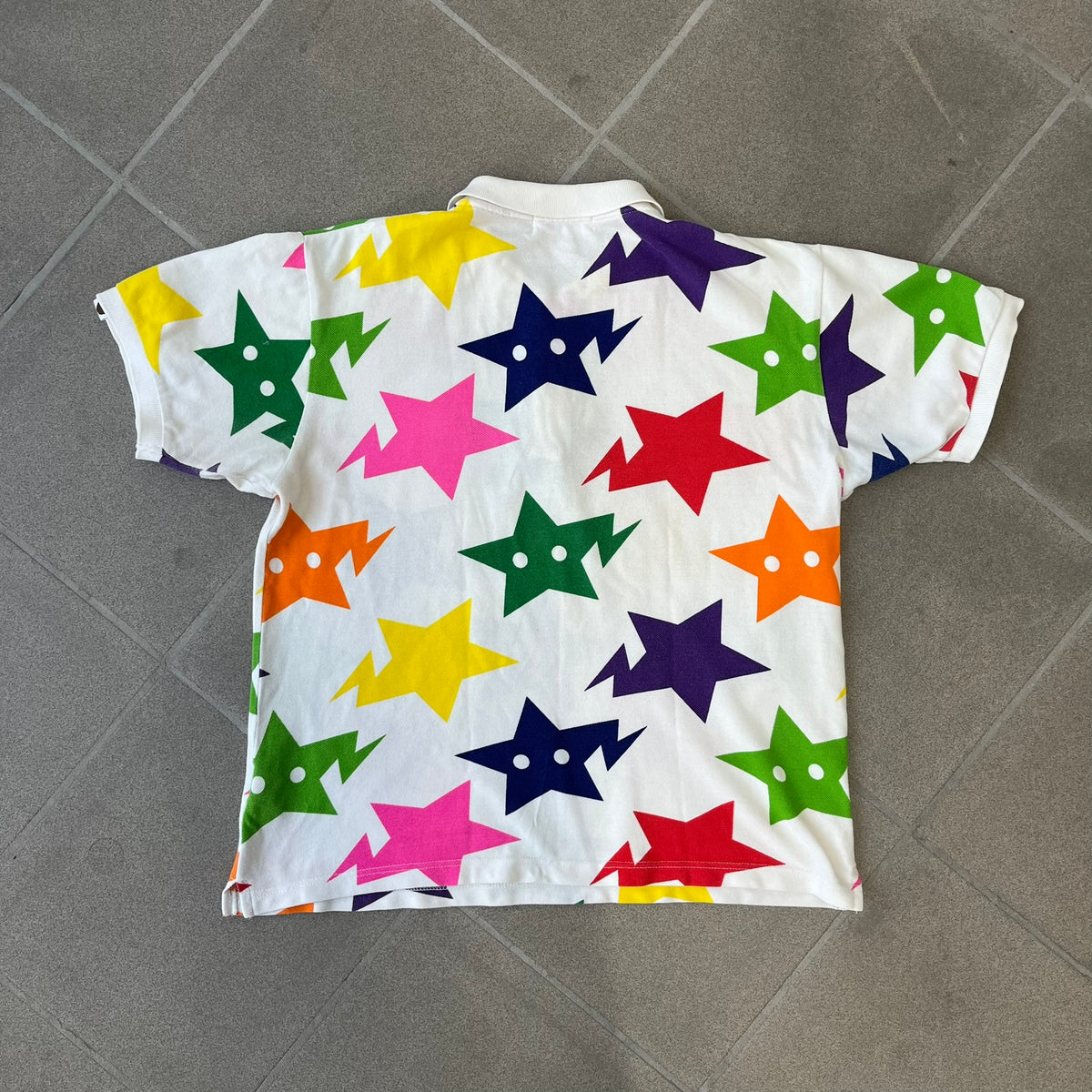 A Bathing Ape 2014 Milo Multicolor Sta Polo is a white, 100% cotton BAPE polo featuring an all-over print of multicolor STA (shooting star) graphics in pink, green, blue, yellow, red (and more), some featuring eyes. It includes branded Bape buttons.