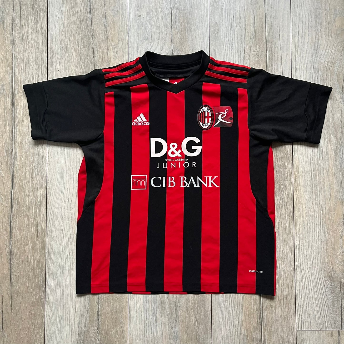 AC Milan x Dolce & Gabbana 2009/10 Home Tee is a black and red striped junior football shirt with black short sleeves. The chest features AC Milan and Adidas logos on the sides, and a D&G Dolce & Gabbana Junior logo in the center. Made with Climacool technology, it is designed for youth performance wear.