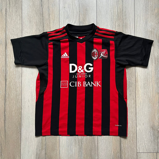 AC Milan x Dolce & Gabbana 2009/10 Home Tee is a black and red striped junior football shirt with black short sleeves. The chest features AC Milan and Adidas logos on the sides, and a D&G Dolce & Gabbana Junior logo in the center. Made with Climacool technology, it is designed for youth performance wear.