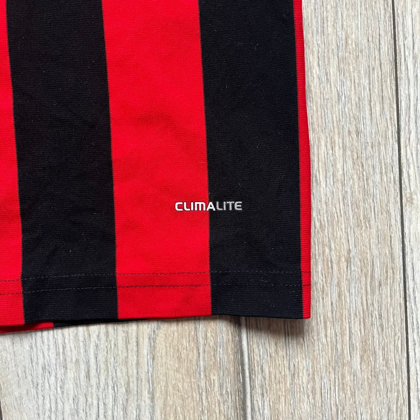 AC Milan x Dolce & Gabbana 2009/10 Home Tee is a black and red striped junior football shirt with black short sleeves. The chest features AC Milan and Adidas logos on the sides, and a D&G Dolce & Gabbana Junior logo in the center. Made with Climacool technology, it is designed for youth performance wear.