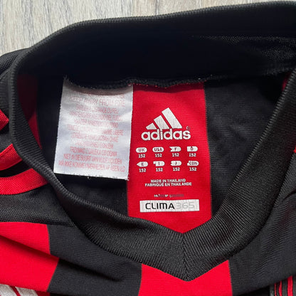 AC Milan x Dolce & Gabbana 2009/10 Home Tee is a black and red striped junior football shirt with black short sleeves. The chest features AC Milan and Adidas logos on the sides, and a D&G Dolce & Gabbana Junior logo in the center. Made with Climacool technology, it is designed for youth performance wear.