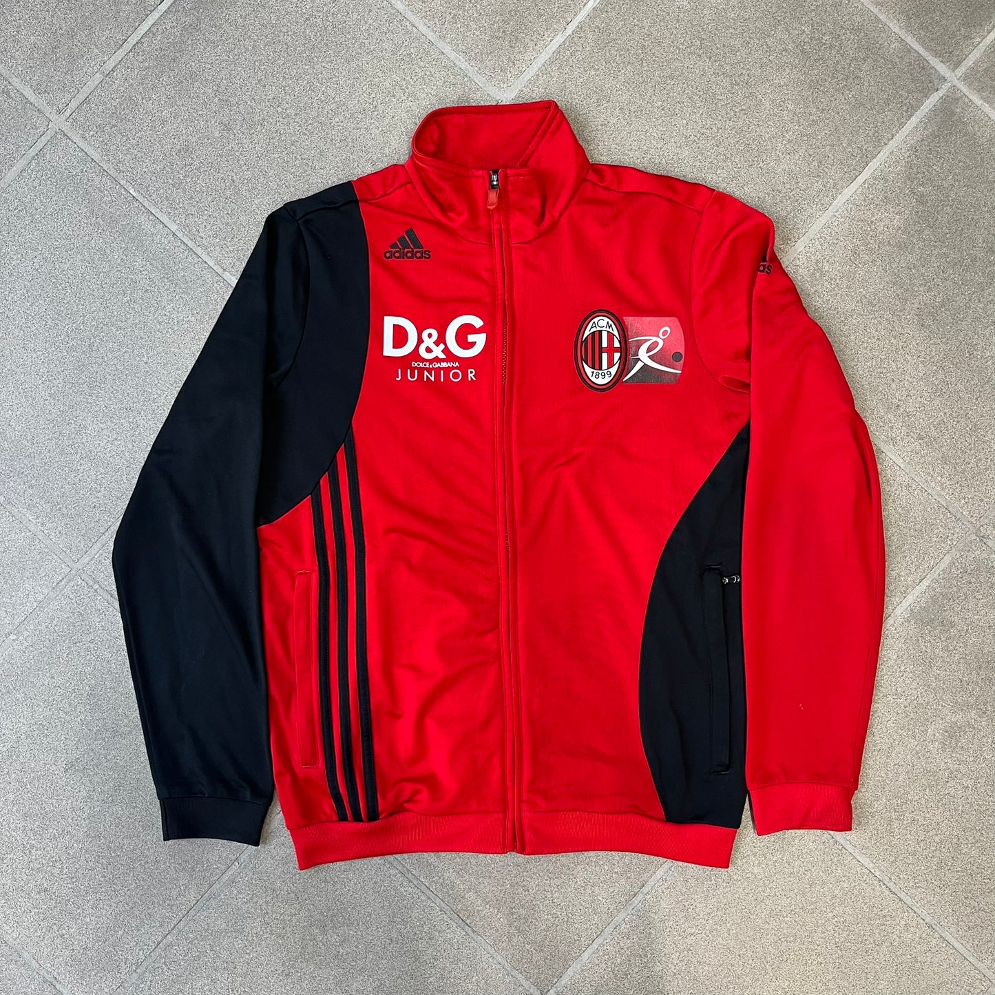 AC Milan x Dolce &amp; Gabbana 2009/10 Track Jacket is a black and red junior football track jacket with asymetrical black and red pattern. The chest features Adidas and D&amp;G Dolce &amp; Gabbana Junior logos on one side, and a AC Milan logo on the left. Made with Climacool technology, it is designed for youth performance wear.