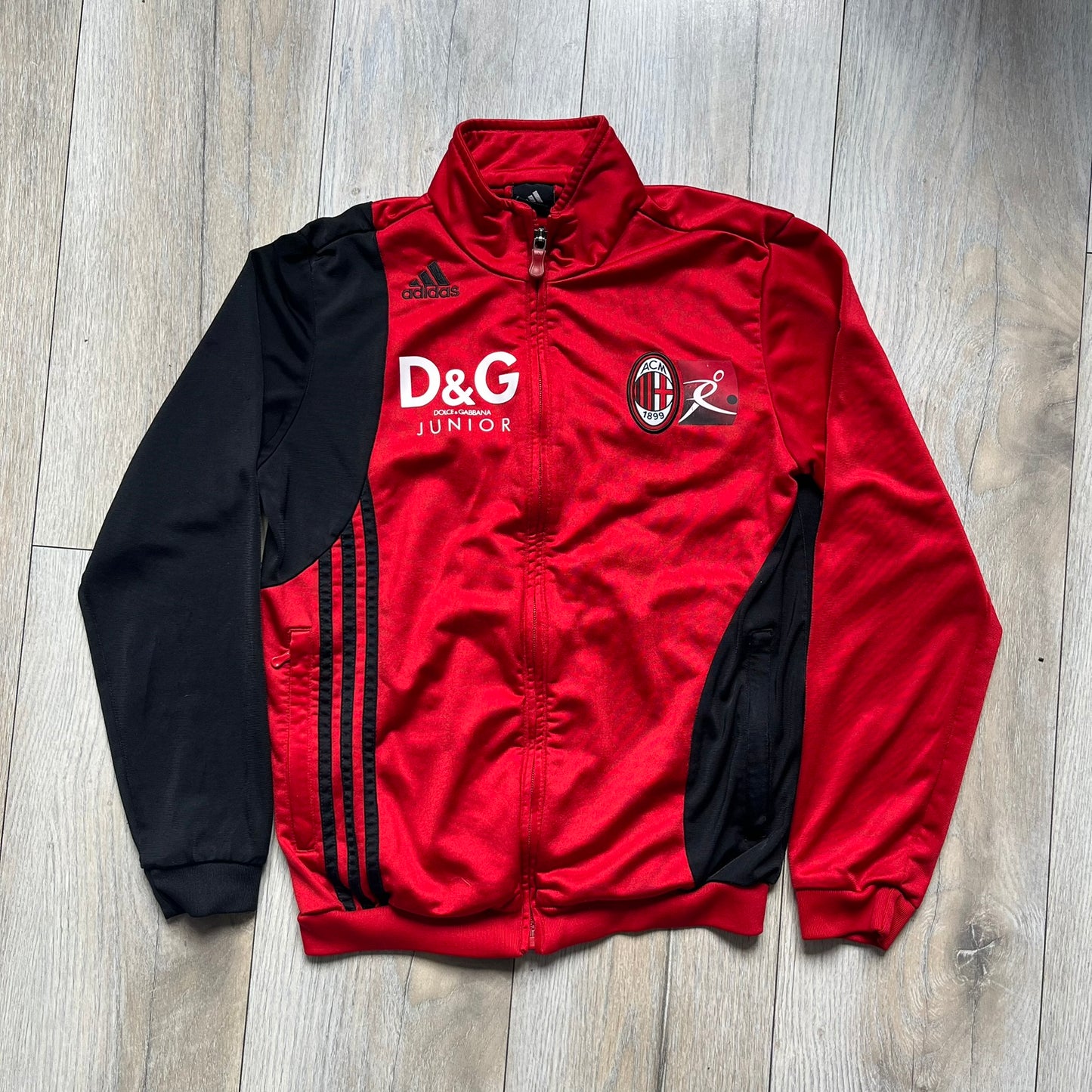 AC Milan x Dolce &amp; Gabbana 2009/10 Track Jacket is a black and red junior football track jacket with asymetrical black and red pattern. The chest features AC Milan and Adidas logos on one side, and a D&amp;G Dolce &amp; Gabbana Junior logo on the left. Made with Climacool technology, it is designed for youth performance wear.