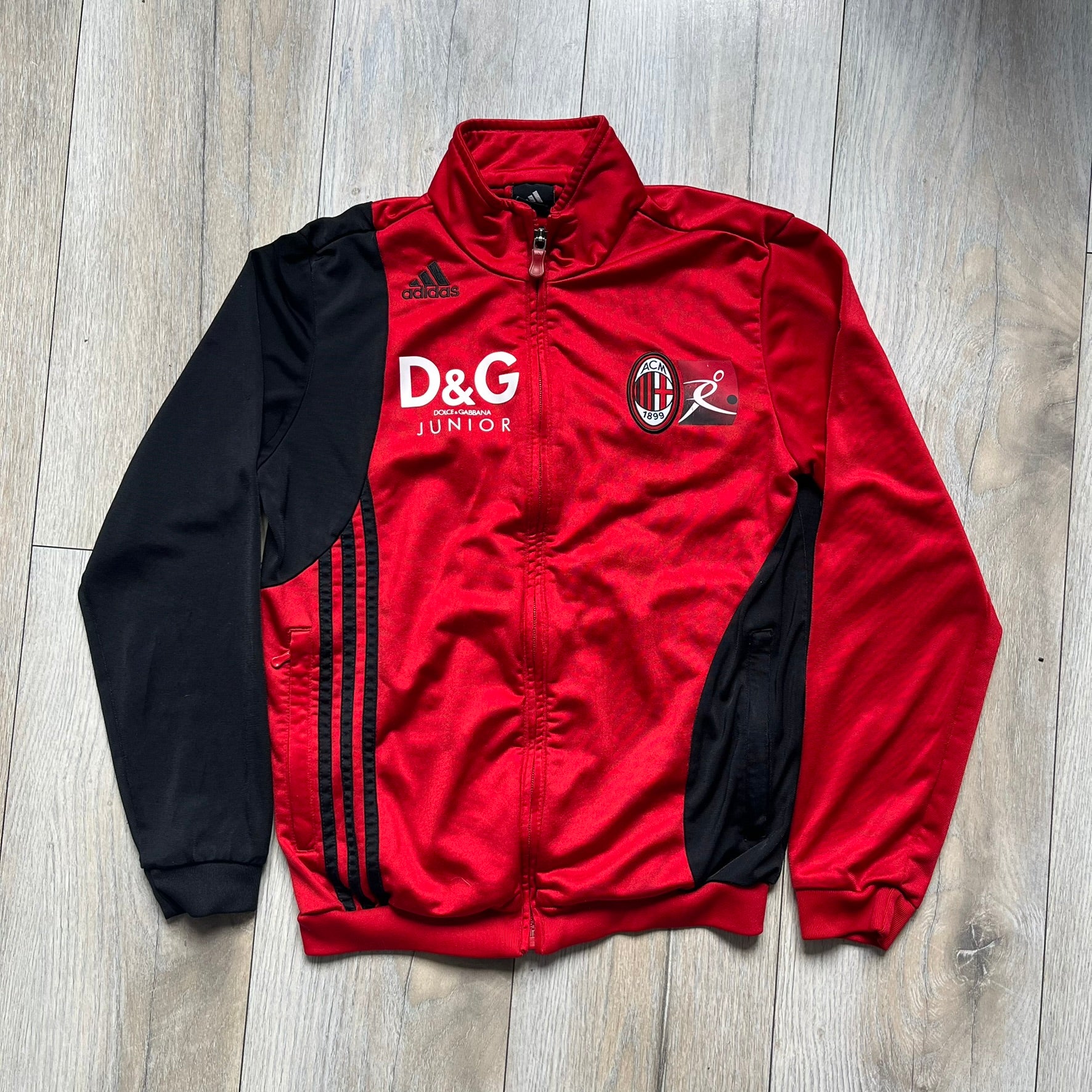 AC Milan x Dolce &amp; Gabbana 2009/10 Track Jacket is a black and red junior football track jacket with asymetrical black and red pattern. The chest features AC Milan and Adidas logos on one side, and a D&amp;G Dolce &amp; Gabbana Junior logo on the left. Made with Climacool technology, it is designed for youth performance wear.