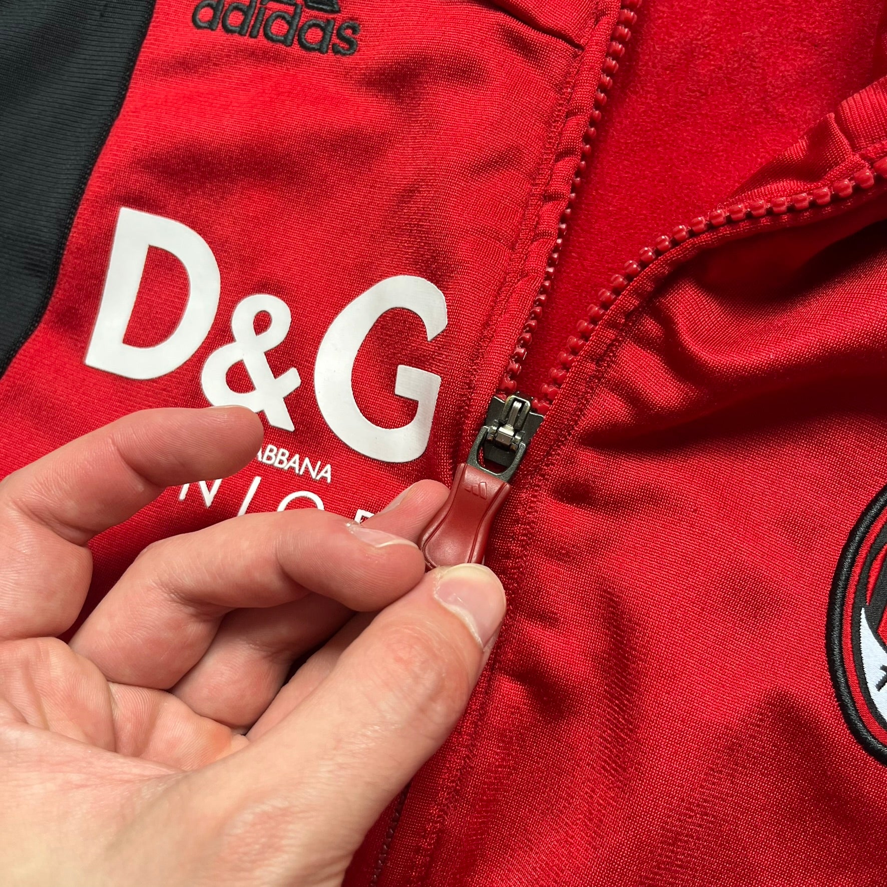 AC Milan x Dolce &amp; Gabbana 2009/10 Track Jacket is a black and red junior football track jacket with asymetrical black and red pattern. The chest features AC Milan and Adidas logos on one side, and a D&amp;G Dolce &amp; Gabbana Junior logo on the left. Made with Climacool technology, it is designed for youth performance wear.