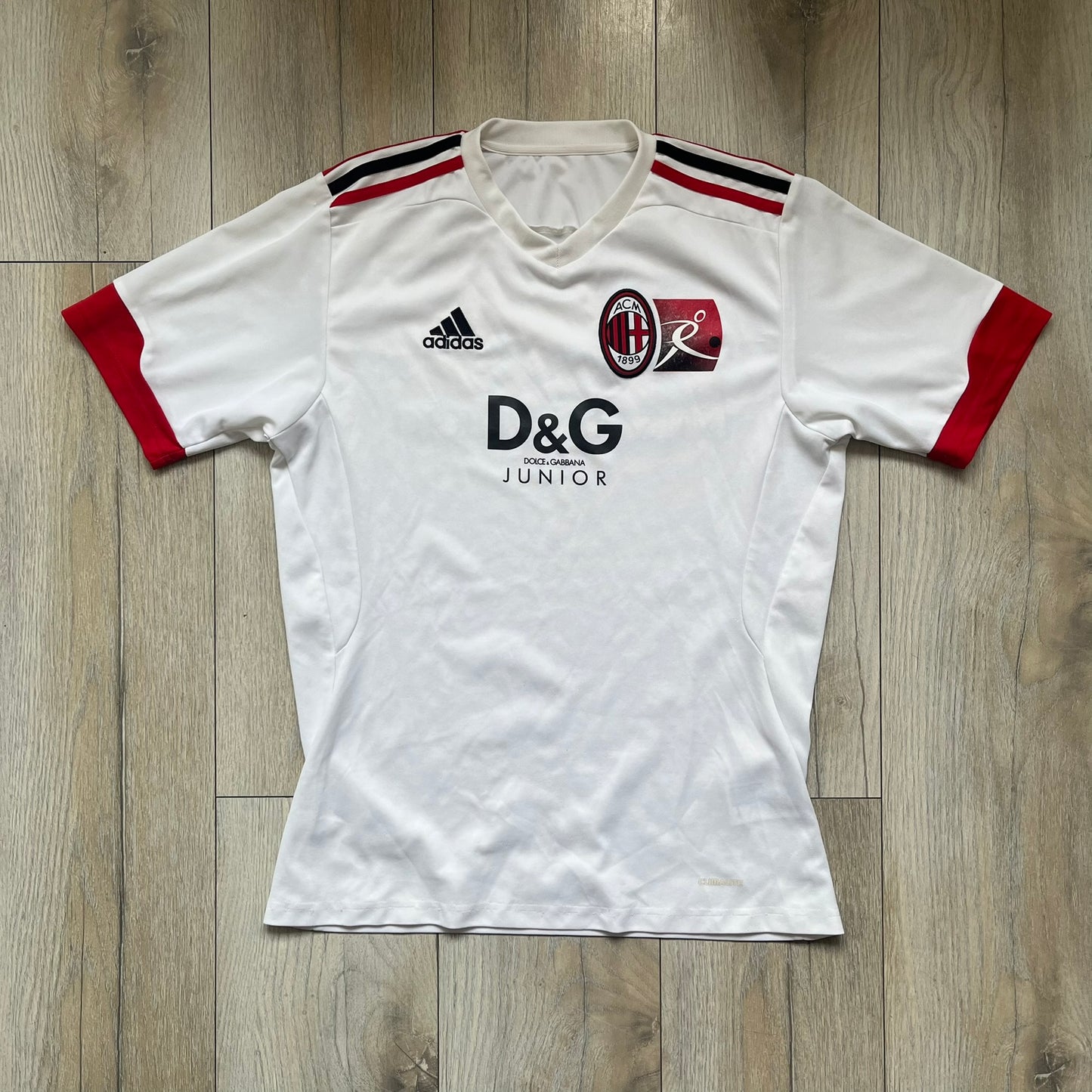 AC Milan x Dolce &amp; Gabbana 2009/10 Training Tee is a white football shirt with red ended short sleeves. The chest features AC Milan and Adidas logos on the sides, and a D&amp;G Dolce &amp; Gabbana Junior logo in the center. Made with Climacool technology, it is designed for performance wear.
