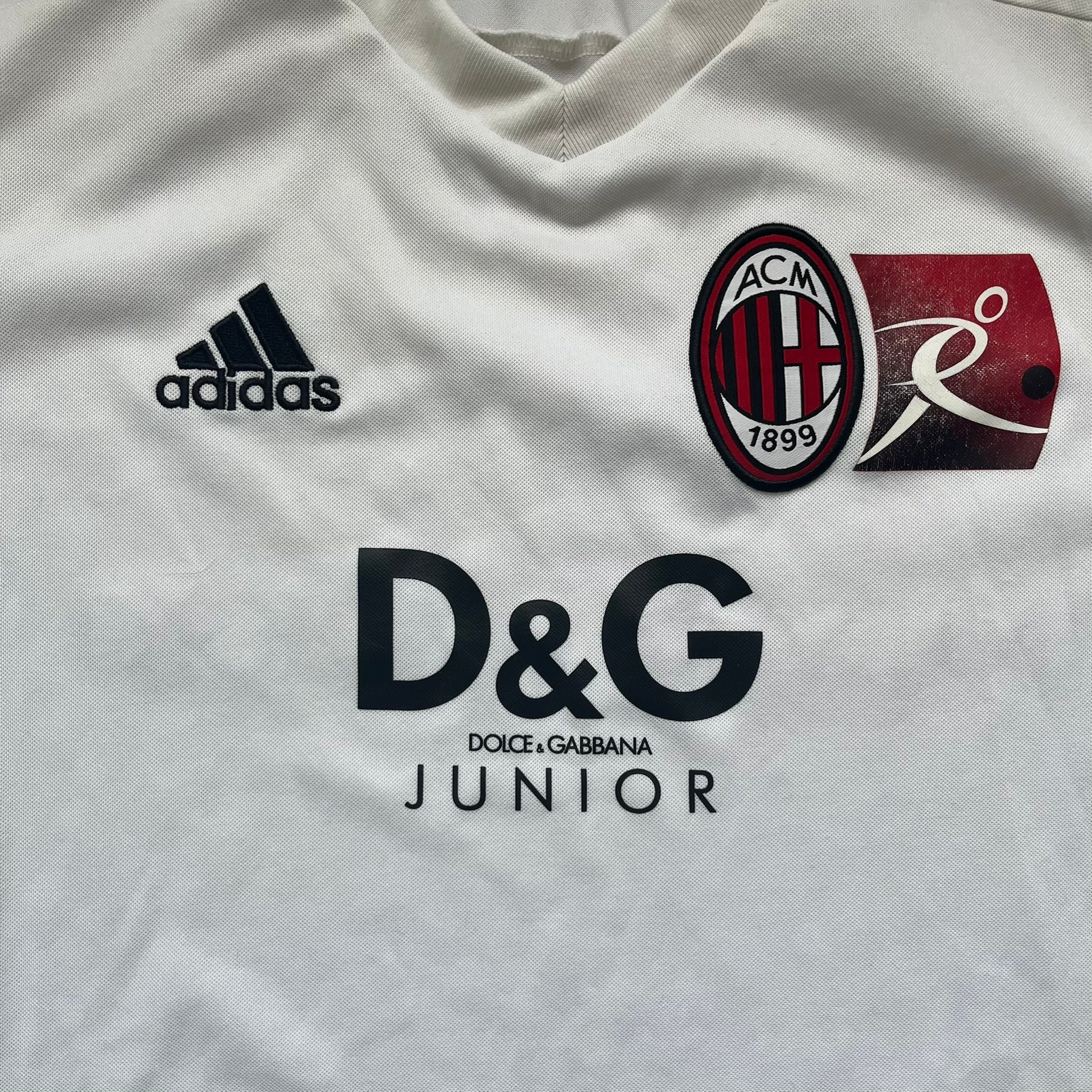 AC Milan x Dolce &amp; Gabbana 2009/10 Training Tee is a white football shirt with red ended short sleeves. The chest features AC Milan and Adidas logos on the sides, and a D&amp;G Dolce &amp; Gabbana Junior logo in the center. Made with Climacool technology, it is designed for performance wear.
