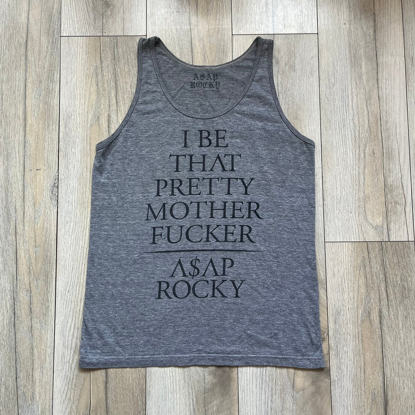 A$AP Rocky 2012 “Peso” Gray Tank Top is a grey sleeveless shirt sold at concerts during the early A$AP Rocky tours. The front features stacked text reading “I be that pretty mother fucker” arranged word-by-word from top to bottom, followed by a line and “ASAP Rocky” beneath. The neckline includes A$AP Rocky branding printed on the tag area.