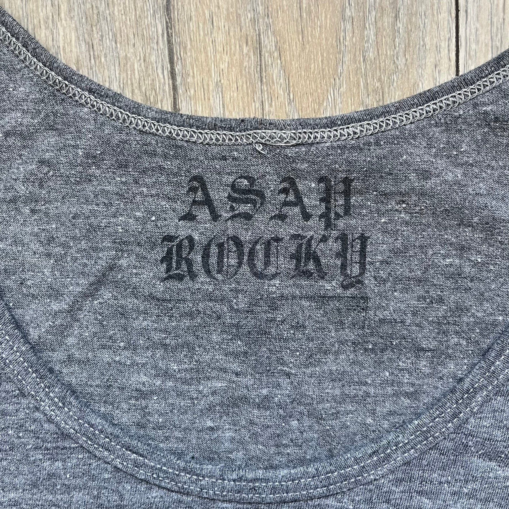 A$AP Rocky 2012 “Peso” Gray Tank Top is a grey sleeveless shirt sold at concerts during the early A$AP Rocky tours. The front features stacked text reading “I be that pretty mother fucker” arranged word-by-word from top to bottom, followed by a line and “ASAP Rocky” beneath. The neckline includes A$AP Rocky branding printed on the tag area.