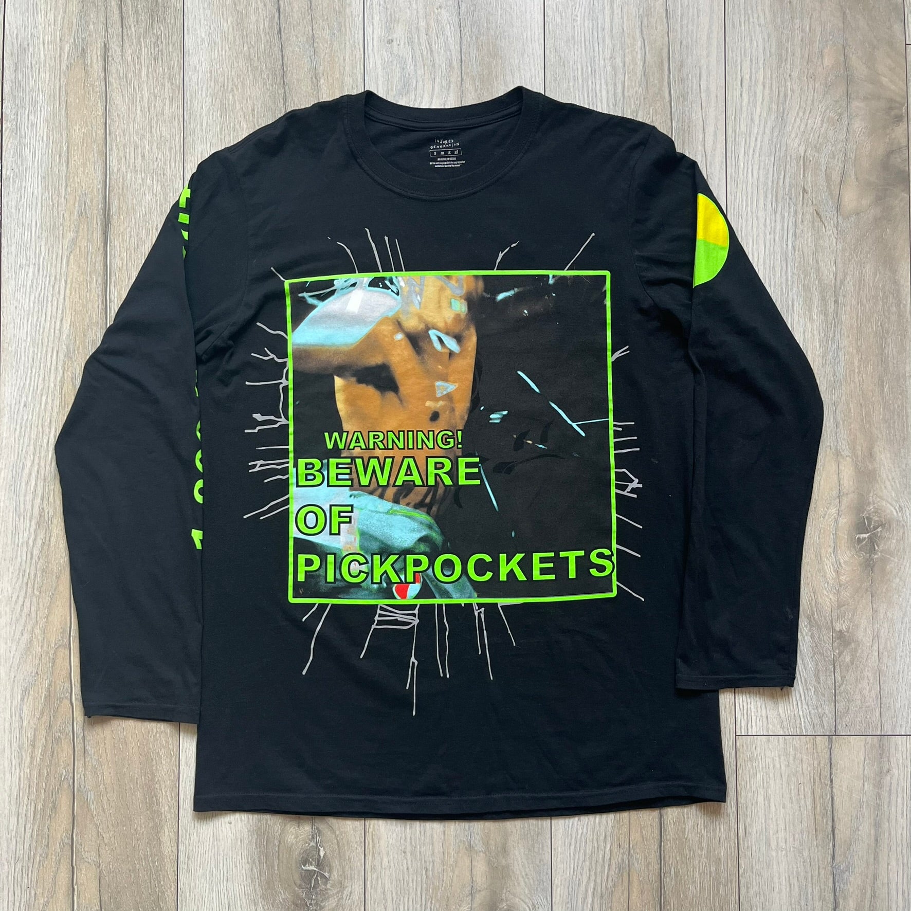 A$AP Rocky 2019 “Injured Generation” Tour “Pickpockets” Longsleeve is a black long-sleeve T-shirt released as part of the Injured Generation tour merchandise collection. The front features a large graphic of A$AP Rocky with green “Warning! Beware of Pickpockets” text. One sleeve includes “1-800-EAT-SHIT” text, while the other features a yellow and green testing-style logo. The back displays large green “Don’t Be a Dummy A$AP Rocky Europe” text layered over a white dummy mask graphic. Made in the USA.