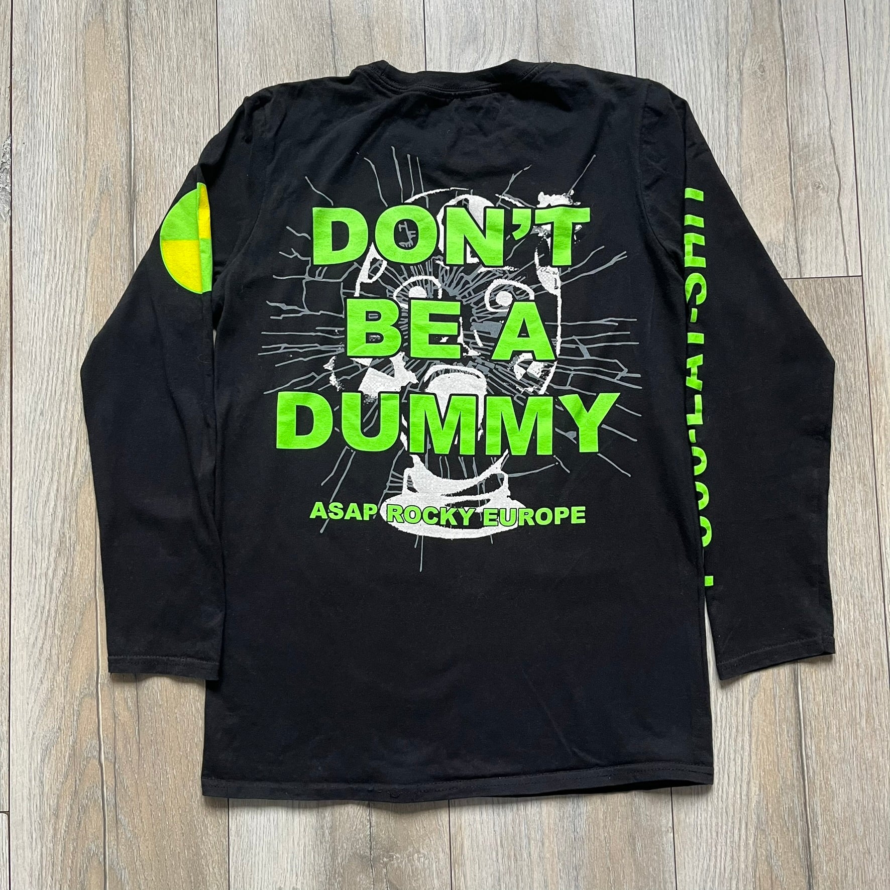 A$AP Rocky 2019 “Injured Generation” Tour “Pickpockets” Longsleeve is a black long-sleeve T-shirt released as part of the Injured Generation tour merchandise collection. The front features a large graphic of A$AP Rocky with green “Warning! Beware of Pickpockets” text. One sleeve includes “1-800-EAT-SHIT” text, while the other features a yellow and green testing-style logo. The back displays large green “Don’t Be a Dummy A$AP Rocky Europe” text layered over a white dummy mask graphic. Made in the USA.