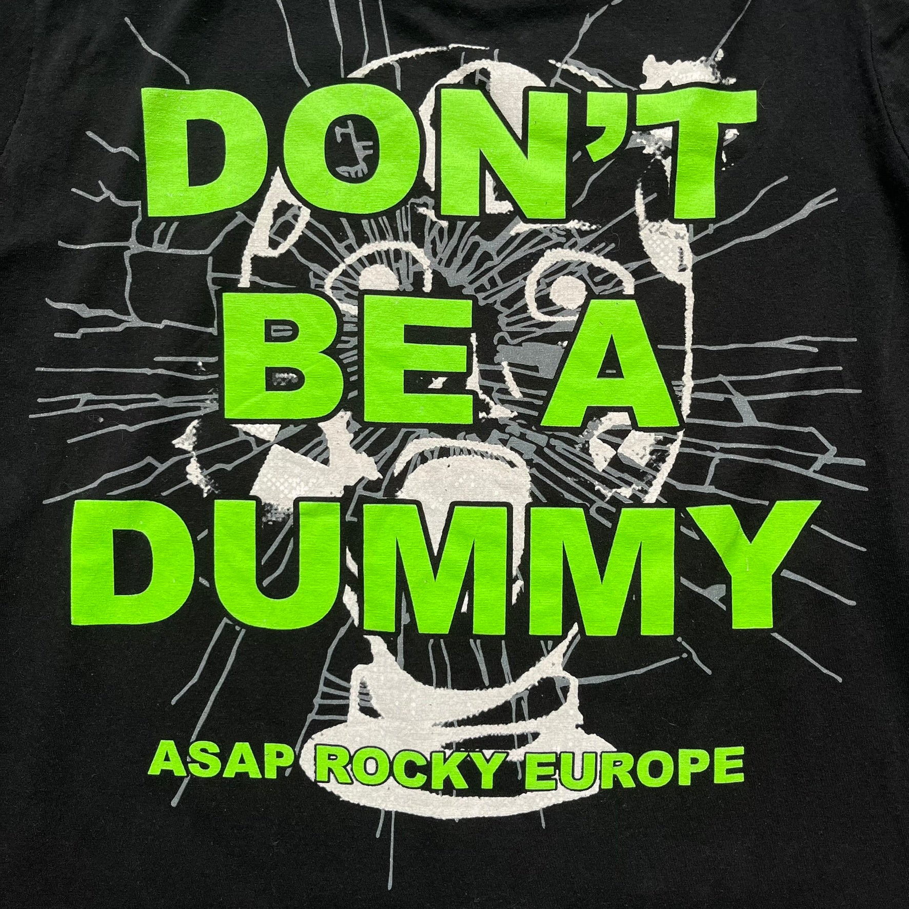 A$AP Rocky 2019 “Injured Generation” Tour “Pickpockets” Longsleeve is a black long-sleeve T-shirt released as part of the Injured Generation tour merchandise collection. The front features a large graphic of A$AP Rocky with green “Warning! Beware of Pickpockets” text. One sleeve includes “1-800-EAT-SHIT” text, while the other features a yellow and green testing-style logo. The back displays large green “Don’t Be a Dummy A$AP Rocky Europe” text layered over a white dummy mask graphic. Made in the USA.