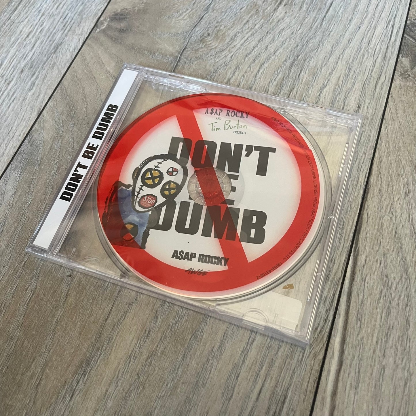 A$AP Rocky “Don’t Be Dumb” GRIM CD is a jewel case CD release of Don’t Be Dumb by A$AP Rocky featuring DUMMY artwork by Tim Burton. The cover includes large “Don’t Be Dumb” text with smaller “A$AP Rocky” and “Tim Burton Presents” above, along with the Dummy figure in a Dummy mask. The case spine features “Don’t Be Dumb” text, and the disc includes a foil design with a red crossed-out circle graphic.