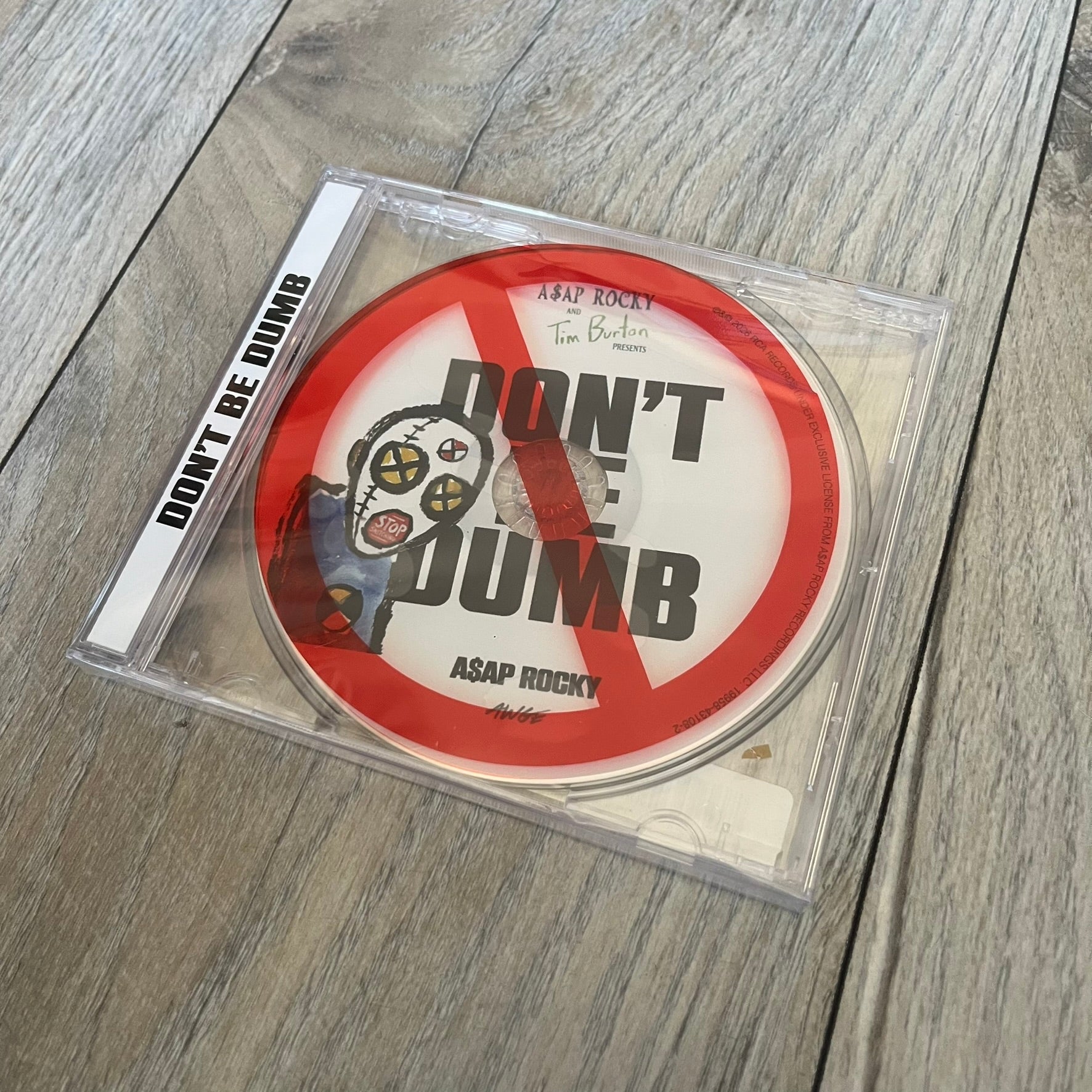 A$AP Rocky “Don’t Be Dumb” GRIM CD is a jewel case CD release of Don’t Be Dumb by A$AP Rocky featuring DUMMY artwork by Tim Burton. The cover includes large “Don’t Be Dumb” text with smaller “A$AP Rocky” and “Tim Burton Presents” above, along with the Dummy figure in a Dummy mask. The case spine features “Don’t Be Dumb” text, and the disc includes a foil design with a red crossed-out circle graphic.