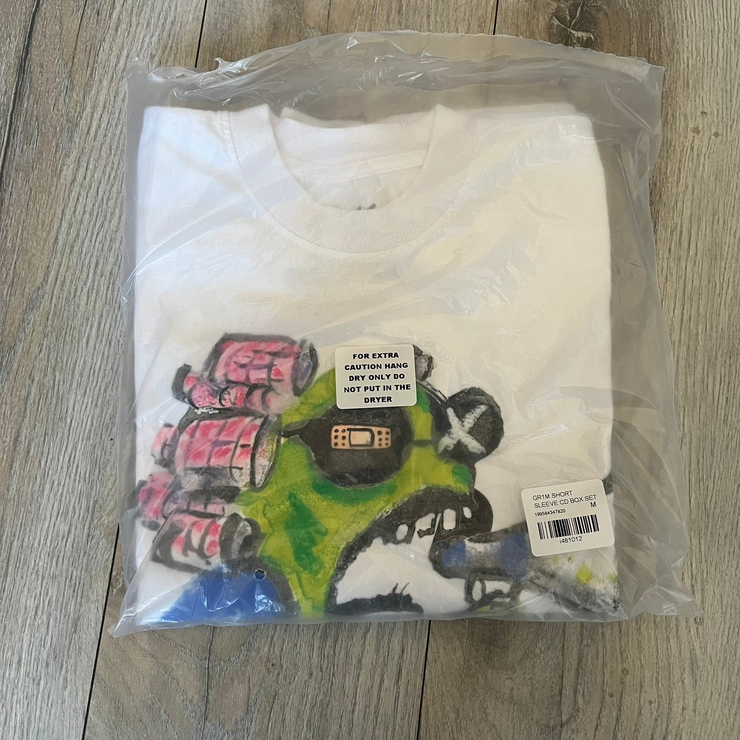 A$AP Rocky “Don’t Be Dumb” GR1M Short Sleeve Tee is a limited release from the AWGE website tied to the rollout of Don’t Be Dumb by A$AP Rocky. The white T-shirt features a green GR1M figure with a megaphone printed on the front with “GRIM” text below. The reverse includes a framed graphic with “Don’t Be Dumb” text, using a lenticular-style vinyl panel that shifts between the GR1M figure and A$AP Rocky in the same pose.