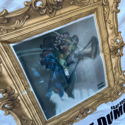 A$AP Rocky “Don’t Be Dumb” GR1M Short Sleeve Tee is a limited release from the AWGE website tied to the rollout of Don’t Be Dumb by A$AP Rocky. The white T-shirt features a green GR1M figure with a megaphone printed on the front with “GRIM” text below. The reverse includes a framed graphic with “Don’t Be Dumb” text, using a lenticular-style vinyl panel that shifts between the GR1M figure and A$AP Rocky in the same pose.
