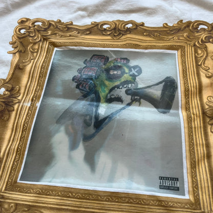 A$AP Rocky “Don’t Be Dumb” GR1M Short Sleeve Tee is a limited release from the AWGE website tied to the rollout of Don’t Be Dumb by A$AP Rocky. The white T-shirt features a green GR1M figure with a megaphone printed on the front with “GRIM” text below. The reverse includes a framed graphic with “Don’t Be Dumb” text, using a lenticular-style vinyl panel that shifts between the GR1M figure and A$AP Rocky in the same pose.