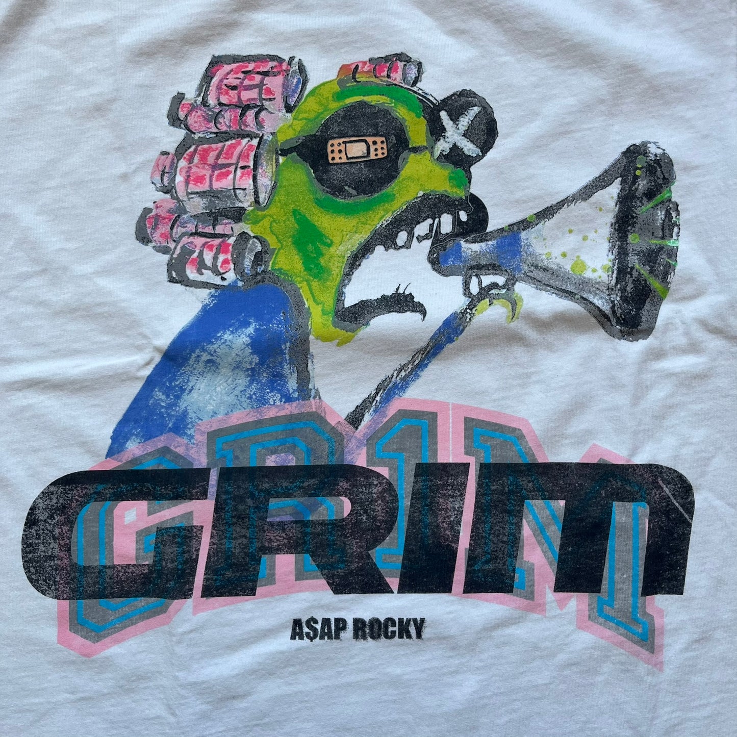 A$AP Rocky “Don’t Be Dumb” GR1M Short Sleeve Tee is a limited release from the AWGE website tied to the rollout of Don’t Be Dumb by A$AP Rocky. The white T-shirt features a green GR1M figure with a megaphone printed on the front with “GRIM” text below. The reverse includes a framed graphic with “Don’t Be Dumb” text, using a lenticular-style vinyl panel that shifts between the GR1M figure and A$AP Rocky in the same pose.