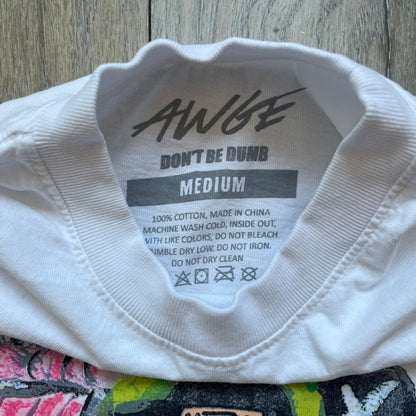 A$AP Rocky “Don’t Be Dumb” GR1M Short Sleeve Tee is a limited release from the AWGE website tied to the rollout of Don’t Be Dumb by A$AP Rocky. The white T-shirt features a green GR1M figure with a megaphone printed on the front with “GRIM” text below. The reverse includes a framed graphic with “Don’t Be Dumb” text, using a lenticular-style vinyl panel that shifts between the GR1M figure and A$AP Rocky in the same pose.