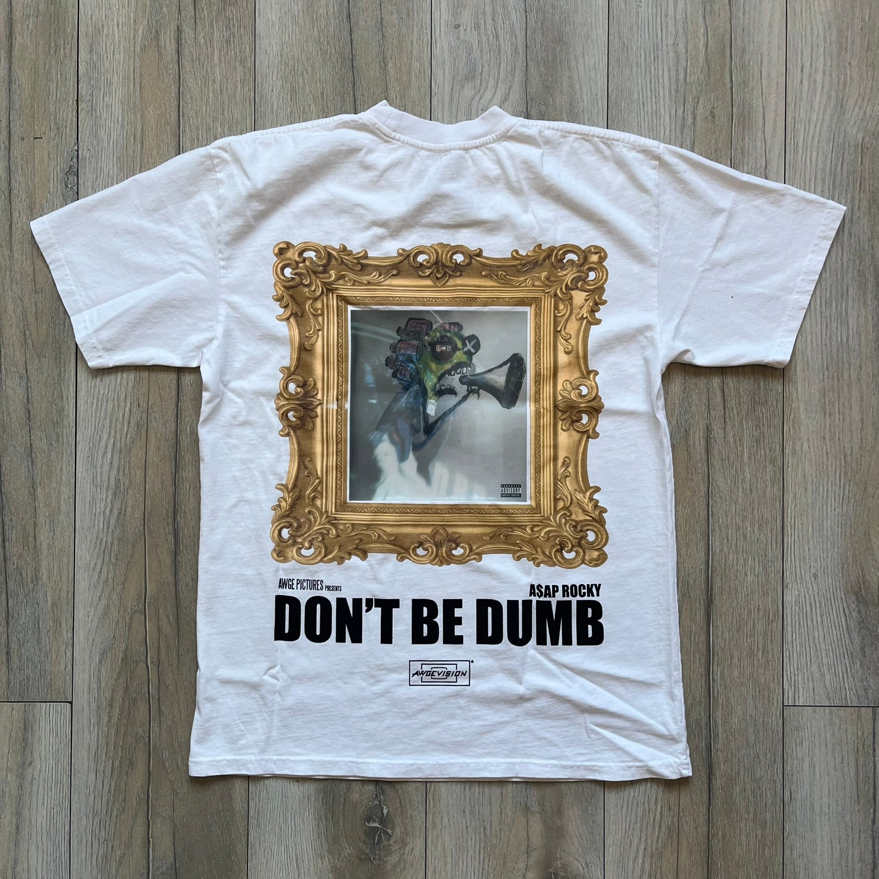 A$AP Rocky “Don’t Be Dumb” GR1M Short Sleeve Tee is a limited release from the AWGE website tied to the rollout of Don’t Be Dumb by A$AP Rocky. The white T-shirt features a green GR1M figure with a megaphone printed on the front with “GRIM” text below. The reverse includes a framed graphic with “Don’t Be Dumb” text, using a lenticular-style vinyl panel that shifts between the GR1M figure and A$AP Rocky in the same pose.