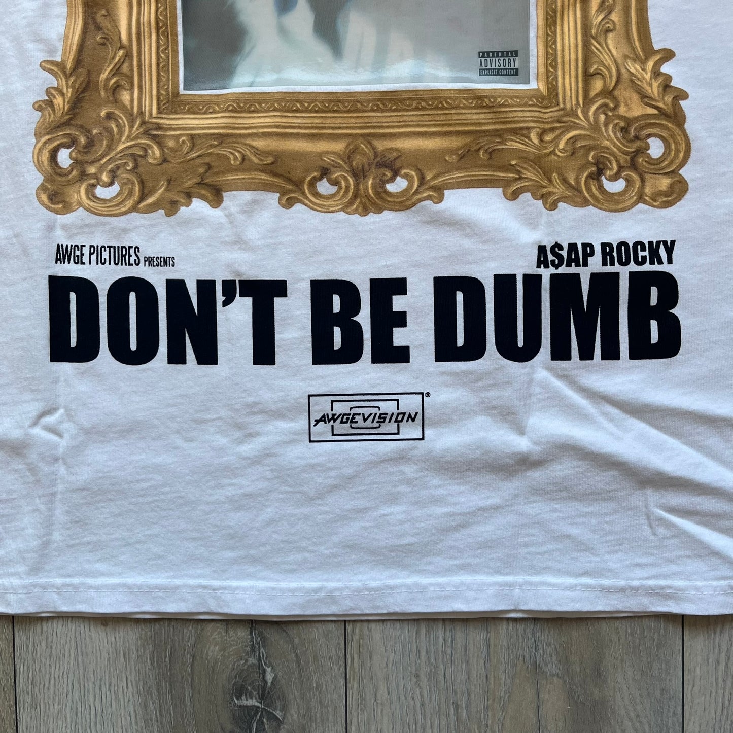 A$AP Rocky “Don’t Be Dumb” GR1M Short Sleeve Tee is a limited release from the AWGE website tied to the rollout of Don’t Be Dumb by A$AP Rocky. The white T-shirt features a green GR1M figure with a megaphone printed on the front with “GRIM” text below. The reverse includes a framed graphic with “Don’t Be Dumb” text, using a lenticular-style vinyl panel that shifts between the GR1M figure and A$AP Rocky in the same pose.