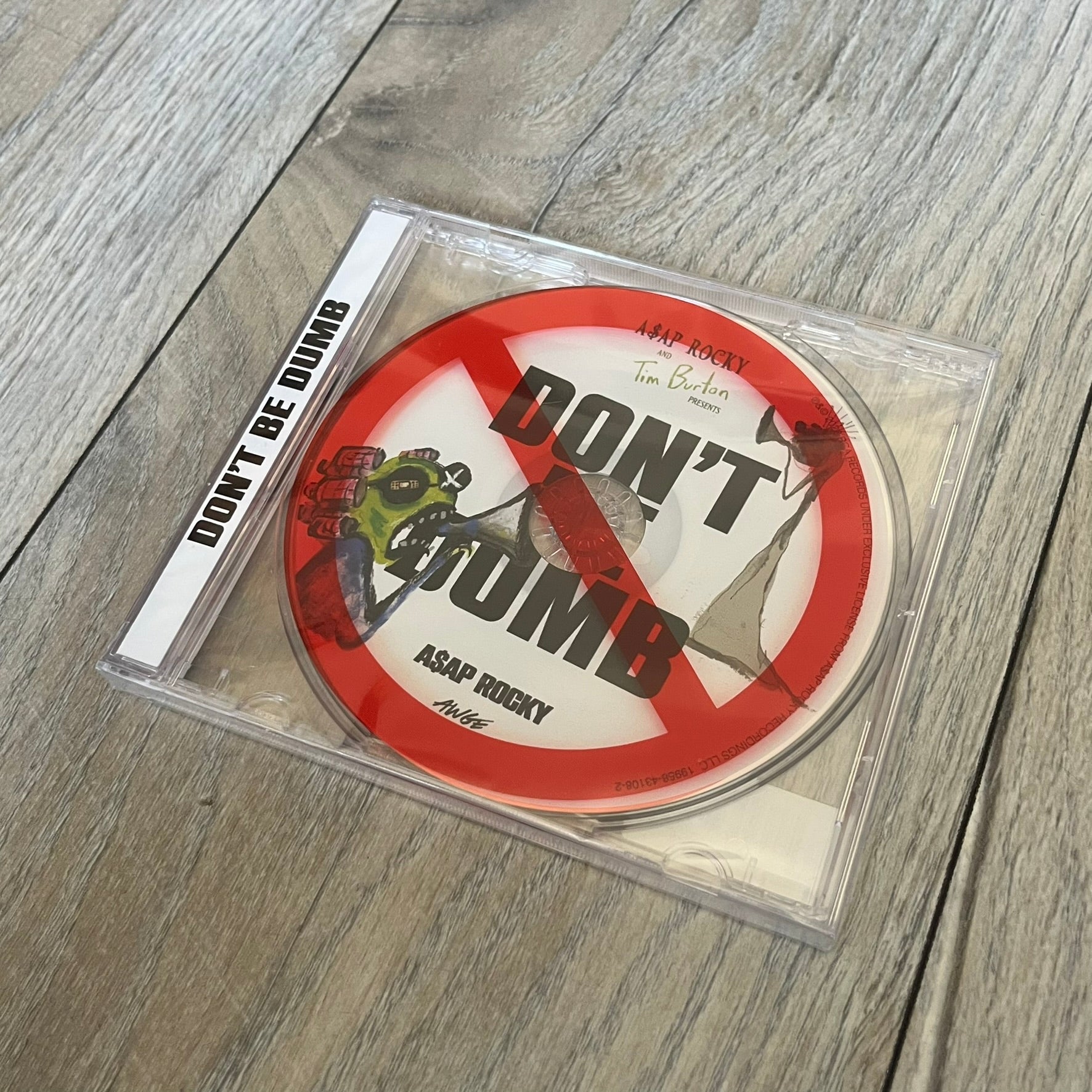 A$AP Rocky “Don’t Be Dumb” GRIM CD is a jewel case CD release of Don’t Be Dumb by A$AP Rocky featuring GRIM artwork by Tim Burton. The cover includes large “Don’t Be Dumb” text with smaller “A$AP Rocky” and “Tim Burton Presents” above, along with the GRIM figure holding a megaphone. The case spine features “Don’t Be Dumb” text, and the disc includes a foil design with a red crossed-out circle graphic.