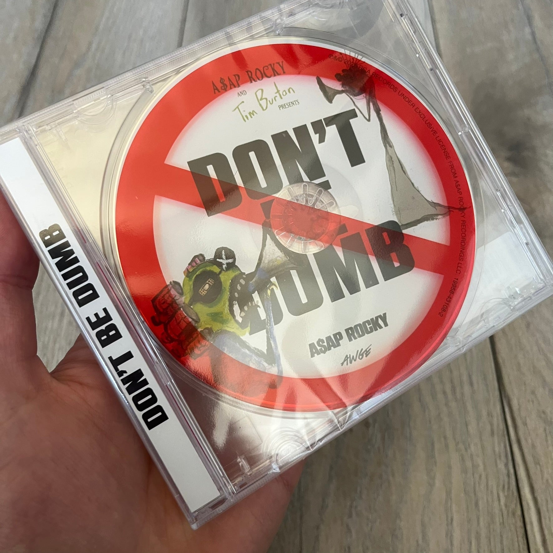 A$AP Rocky “Don’t Be Dumb” GRIM CD is a jewel case CD release of Don’t Be Dumb by A$AP Rocky featuring GRIM artwork by Tim Burton. The cover includes large “Don’t Be Dumb” text with smaller “A$AP Rocky” and “Tim Burton Presents” above, along with the GRIM figure holding a megaphone. The case spine features “Don’t Be Dumb” text, and the disc includes a foil design with a red crossed-out circle graphic.