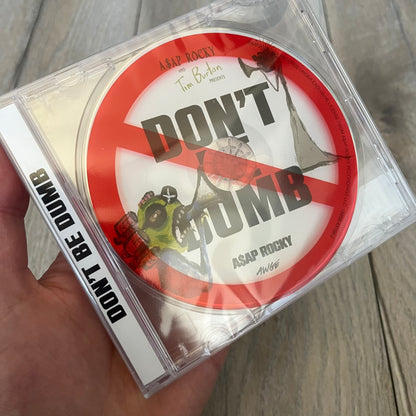 A$AP Rocky “Don’t Be Dumb” GRIM CD is a jewel case CD release of Don’t Be Dumb by A$AP Rocky featuring GRIM artwork by Tim Burton. The cover includes large “Don’t Be Dumb” text with smaller “A$AP Rocky” and “Tim Burton Presents” above, along with the GRIM figure holding a megaphone. The case spine features “Don’t Be Dumb” text, and the disc includes a foil design with a red crossed-out circle graphic.