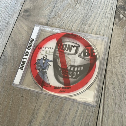 A$AP Rocky “Don’t Be Dumb” GRIM CD is a jewel case CD release of Don’t Be Dumb by A$AP Rocky featuring Mr. Mayers artwork by Tim Burton. The cover includes large “Don’t Be Dumb” text with smaller “A$AP Rocky” and “Tim Burton Presents” above, along with the Mr. Mayers figure in a blue trench coat. The case spine features “Don’t Be Dumb” text, and the disc includes a foil design with a red crossed-out circle graphic.