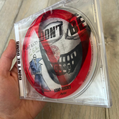 A$AP Rocky “Don’t Be Dumb” GRIM CD is a jewel case CD release of Don’t Be Dumb by A$AP Rocky featuring Mr. Mayers artwork by Tim Burton. The cover includes large “Don’t Be Dumb” text with smaller “A$AP Rocky” and “Tim Burton Presents” above, along with the Mr. Mayers figure in a blue trench coat. The case spine features “Don’t Be Dumb” text, and the disc includes a foil design with a red crossed-out circle graphic.