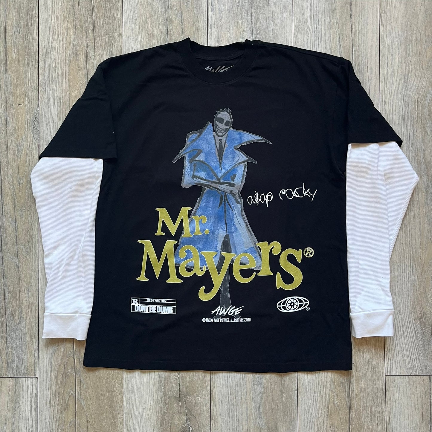 A$AP Rocky “Don’t Be Dumb” Mr Mayers Twofer Longsleeve is a limited release from the AWGE website tied to the rollout of Don’t Be Dumb by A$AP Rocky. The piece features a twofer construction with a black body and white sleeves, with a front graphic of the Mr Mayers figure in a blue trench coat with “Mr Mayers” text and small AWGE logos. The back includes a framed graphic with “Don’t Be Dumb” text, using a lenticular-style vinyl panel that shifts between the Mr Mayers figure and A$AP Rocky in the same outfit
